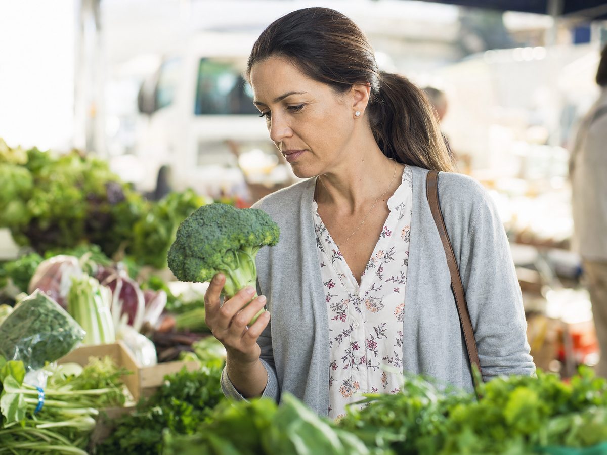 A person reading a supplement label in a bright kitchen with fresh broccoli nearby